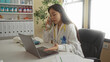 © Krakenimages.com - Woman doctor with stethoscope in clinic working on laptop and talking on phone, surrounded by medical equipment and files in an organized workspace.