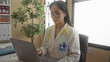 © Krakenimages.com - Serious young chinese woman doctor with stethoscope and uniform crosses arms in a hospital clinic room, focused on a laptop at her workplace.