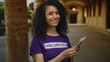 © Krakenimages.com - Woman with curly hair smiling holding dollars wearing purple volunteer shirt outdoors in an old town street with pillars and soft lighting.