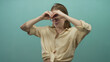 © Krakenimages.com - Young blonde woman makes binoculars gesture in studio with teal background; exploration curiosity playfulness.