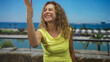 © Krakenimages.com - Woman smiling and raising hand in a pinched fingers gesture, wearing yellow sleeveless top and necklace on a studio seaside set; joyful.