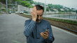 © Krakenimages.com - Hispanic man on seaside promenade checks smartphone with concerned expression, standing near train tracks.