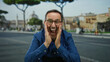 © Krakenimages.com - Hispanic man in blue shirt energetically gesturing outdoors in a city park setting, conveying excitement with hands raised near face against a backdrop of urban architecture.