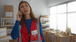 © Krakenimages.com - Volunteer woman talking on phone in charity donations room, wearing a red vest with shelves and packages around, indicating active participation in an indoor support center.