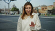 © Krakenimages.com - Young woman in white sweater with uncertain expression stands outdoors on a city street, surrounded by classic architecture and soft lighting, while holding up her hand in a stop gesture.