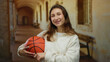 © Krakenimages.com - Woman smiling while holding basketball indoors at an old university hallway with a warm atmosphere reflecting youthful spirit and athleticism for a campus setting.