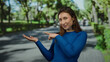 © Krakenimages.com - Young woman in blue sweater pointing and giving thumbs up in a sunny park, surrounded by trees and blurred people.
