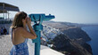 © Krakenimages.com - Woman holding coin operated viewer and looking through blue telescope on street viewpoint overlooking santorini town and caldera; travel exploration joy.