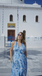 © Krakenimages.com - Woman in blue floral dress holding sunglasses and hand in hair at a white church building in santorini; vacation joy.
