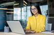 © Tetiana - Smiling and cheerful Indian businesswoman wearing glasses and a yellow shirt working in the office on a laptop