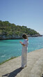 © Krakenimages.com - Woman capturing ocean view on smartphone at sunny beach in santanyi, mallorca with vibrant turquoise water and lush greenery under clear blue sky.