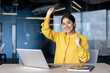 © Tetiana - Portrait of a young Indian woman sitting at a desk in the office, rejoicing and dancing, looking at the camera with a smile