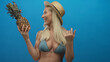 © Krakenimages.com - Woman wearing straw hat and bikini top smiling holds fresh pineapple in a blue studio; tropical happiness.