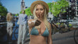 © Krakenimages.com - Blonde woman in bikini top and straw hat displays thumb up and thumb down gesture on street; uncertainty.