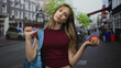 © Krakenimages.com - Young woman holding apple on city street with blue backpack, one arm extended in shrugging pose; confidence casual attitude.
