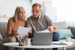 © Prostock-studio - An older couple sits closely on a sofa reviewing important documents. They appear focused and engaged in a discussion. A laptop is open in front of them, adding to the cozy atmosphere.