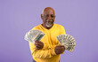 © Prostock-studio - An older man holds several stacks of cash in both hands. He smiles confidently while wearing a yellow sweater against a purple background. The scene feels upbeat and positive.
