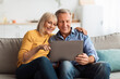 © Prostock-studio - A happy elderly couple sits closely on a sofa, smiling as they watch something on a tablet. Soft natural light fills the warm living room, creating a relaxed atmosphere.