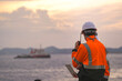© ultramansk - Two engineers wearing safety uniforms and hard hats communicate using a radio while reviewing data on a laptop by the sea during sunset. Maritime industrial setting.