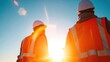 © nan - Two construction workers wearing helmets and high visibility vests stand facing away,