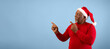 © Prostock-studio - A man wearing a red sweater and a Santa hat stands against a blue background. He points to the left and right as if inviting others to join a festive event during the holiday season.