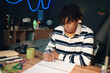 © DragonImages - Black teenage boy sitting at desk writing in notebook with focused expression, surrounded by school supplies in bedroom, studying or doing homework for academic assignment