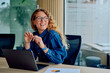 © we.bond.creations - Smiling businesswoman laughing during an office conversation, sitting at a desk with a laptop and documents, expressing joy and confidence