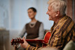 © Seventyfour - Senior Caucasian man playing acoustic guitar while smiling, sitting indoors with blurred middle aged woman in background, nursing home setting suggesting recreational group activity