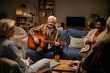 © Seventyfour - Group of seniors including Caucasian man playing acoustic guitar and smiling, senior Caucasian woman and senior Black woman sitting together and listening in nursing home common area