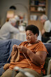 © Seventyfour - Black senior woman sitting on sofa knitting with yarn in nursing home foreground, two Caucasian senior men interacting in background, engaging in leisure activities