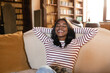 © Prostock-studio - A young woman with long hair smiles joyfully while reclining on a soft couch. Sunlight fills the room, creating a warm and inviting atmosphere. Bookshelves line the walls.