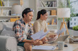 © whyframeshot - Asian married couple sits on their sofa at home, stressfully calculating financial budget and money loan accounts, reflecting unhappiness and debt pressure