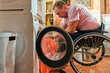 © Alex - A man in a wheelchair removes an orange towel from a washing machine by himself