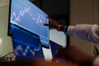 © Studio Marmellata - A person's hand points to a financial chart on a computer screen, analyzing market trends with multiple monitors displaying data.