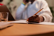 © Studio Marmellata - A person with dark skin writes in a notebook using a pencil on a wooden desk. A glass of water and a laptop are visible, suggesting a focused work or study environment.