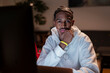 © Studio Marmellata - A young man with braided hair wearing a white hoodie sits at a desk, looking thoughtfully at a computer screen in a dimly lit room.