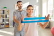 © Studio Romantic - Nurse man helping young female smiling patient to do sport stretching exercises with rubber band in rehabilitation clinic. Male physiotherapist or health care worker helping woman in rehab.