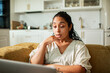 © Marko Geber - Young adult woman working on laptop in home kitchen looking thoughtful