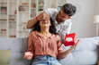 © Home-stock - Indian guy closes eyes to surprised woman and giving present in box, lady sitting on couch in living room. Party at home, birthday gift, anniversary, Valentines day and holiday