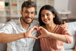 © Home-stock - Affectionate Indian couple smiling and creating heart shape together with their hands, symbolizing love and connection, sitting on sofa at home