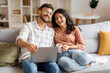 © Home-stock - Excited Indian couple sitting on couch, using laptop and laughing, young man and woman watching movie online, copy space