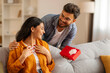 © Home-stock - Surprised Indian woman receiving gift from man standing from back, lady sitting on sofa, both smiling, celebrating anniversary or birthday