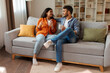 © Home-stock - Loving happy Indian couple sitting on couch in cozy living room, drinking coffee and enjoying pleasant conversation, full length
