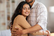 © Home-stock - Affectionate and happy Indian couple hugging, standing in living room interior, woman smiling at camera, free space. Romantic spouses expressing their love