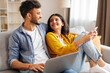 © Home-stock - Positive Indian couple using laptop and smartphone at home, resting on sofa and looking at each other, enjoying weekend pastimes