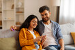 © Home-stock - Banking application and e-commerce. Happy Indian spouses using smartphone and credit card, making online payment in Internet, sitting on sofa at home