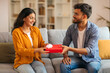 © Home-stock - Loving Indian man giving wrapped gift box to his happy wife, celebrating anniversary or birthday, making surprise, sitting together on sofa at home