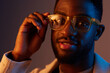 © SHOTPRIME STUDIO - A stylish young African American man adjusting his glasses with a confident smile, illuminated by a warm blue orange background, showcasing modern fashion and self confidence