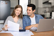 © fizkes - Couple managing family finances in kitchen, sit at table with laptop, calculator, reviewing documents, incomes list, making transaction via e-bank, feel satisfied with earnings growth. Budget, success