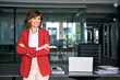 © Stock 4 You - Corporate portrait of mature latin european businesswoman in red blazer standing at desk with laptop in office workplace, arms crossed, looking away, professional female business leader. Copy space